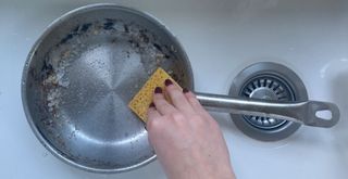 burnt pan in a ceramic sink with a hand using salt and sponge showing how to clean a burnt pan with rock salt