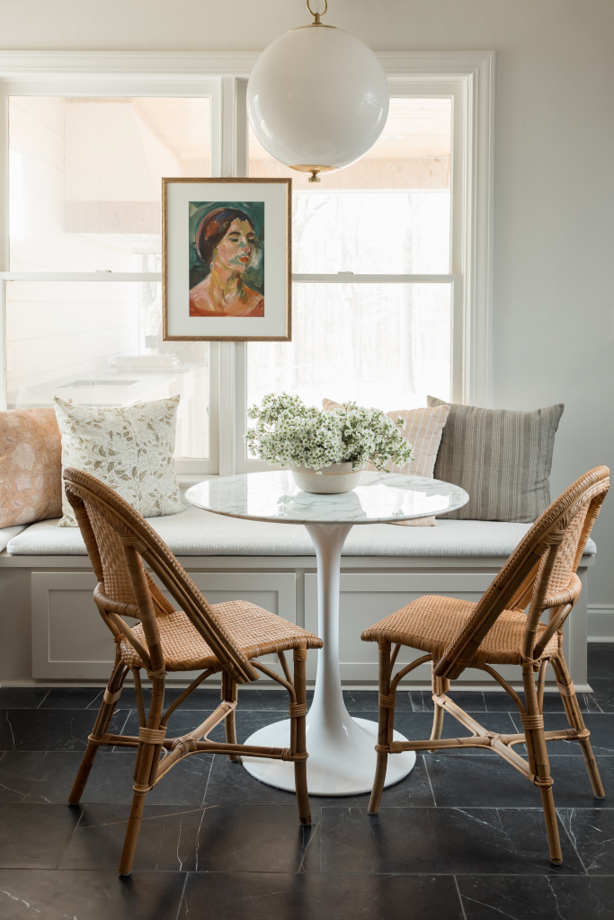 a natural light-filled kitchen featuring a circular table, two wicker chairs, and a pillow-laden bench seat in front of two windows.