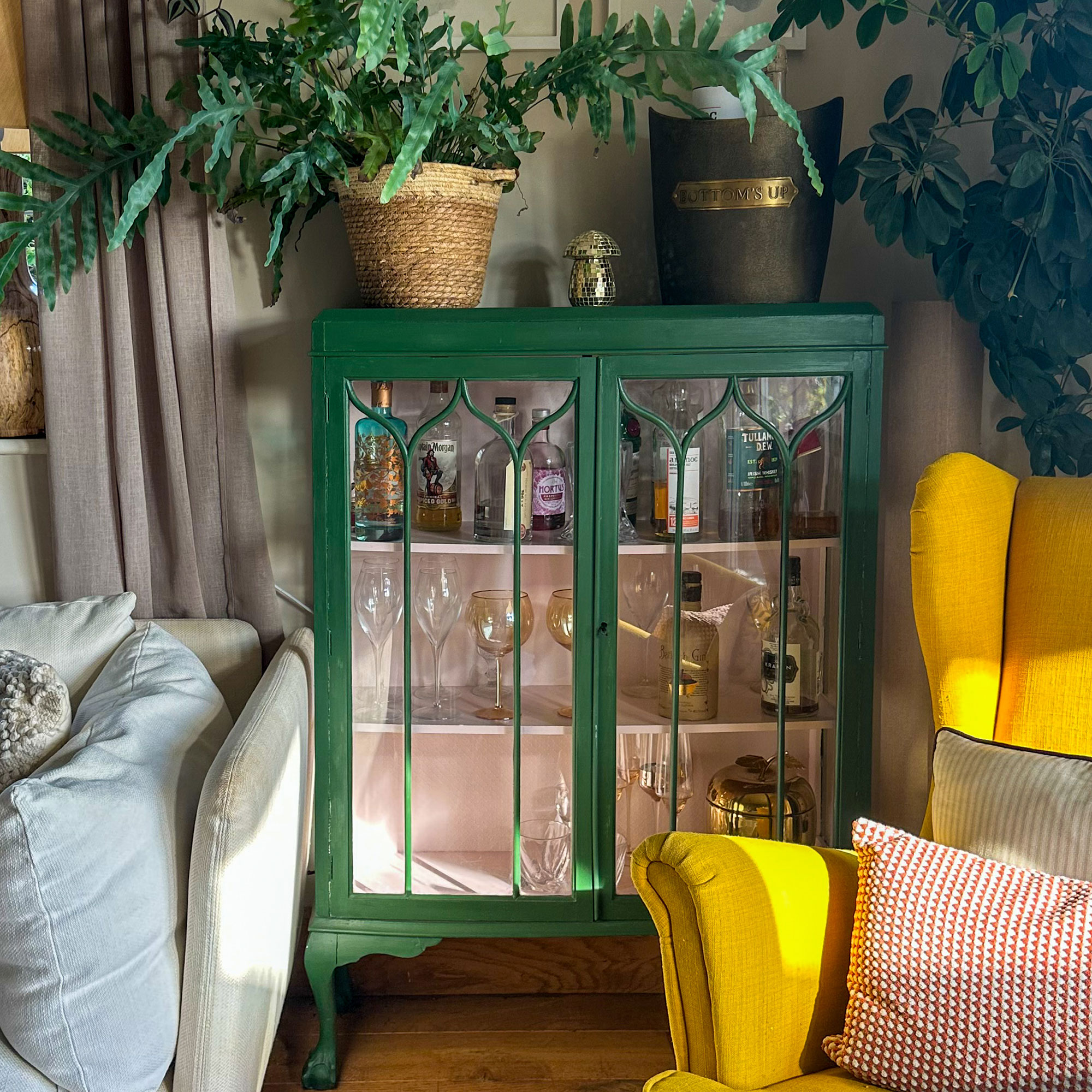 Green glass fronted cabinet holding home bar supplies, tipped with potted plants in living room