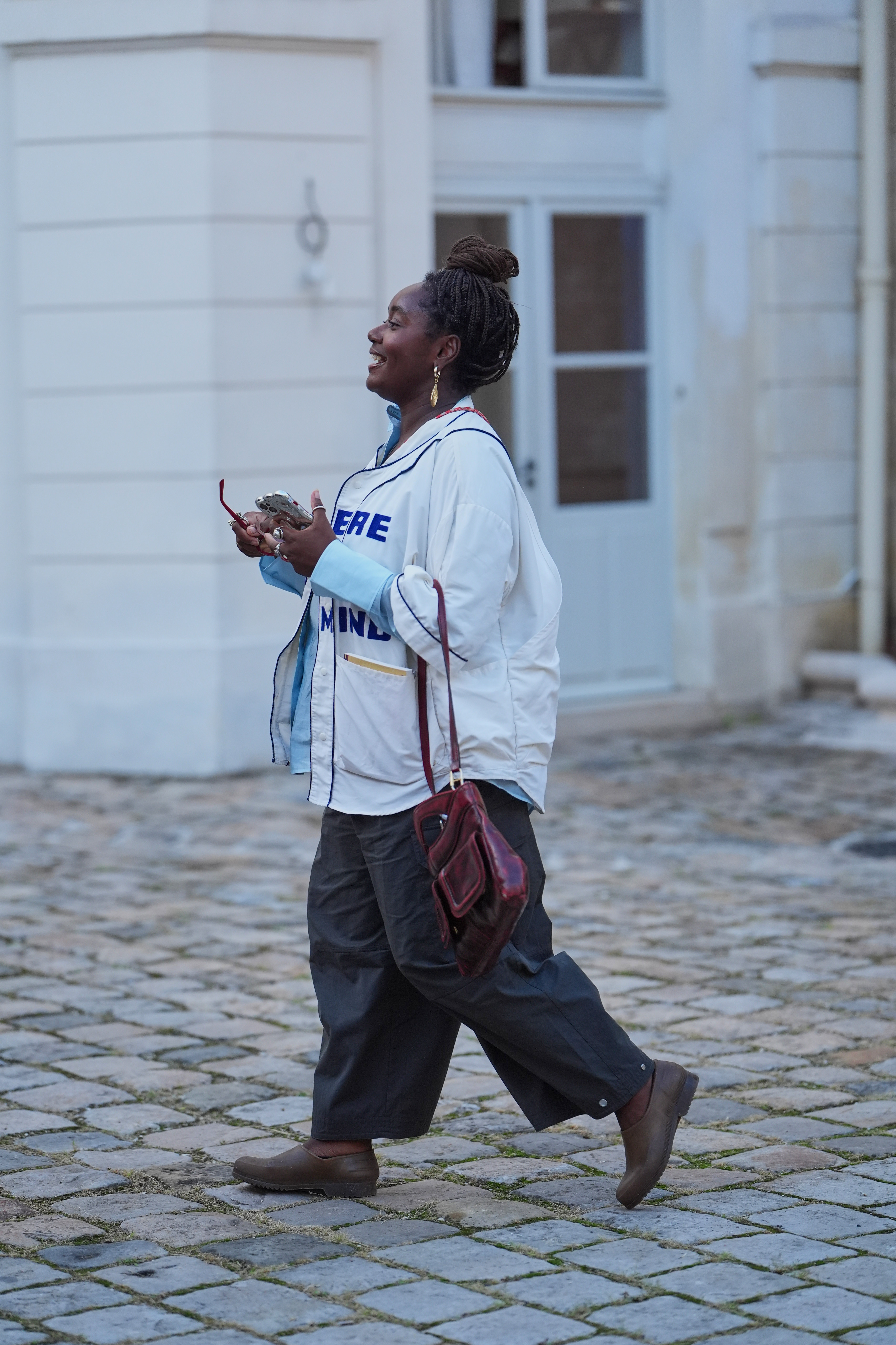 A guest wears a burgundy leather shoulder bag, a white oversized jacket, a light blue button-up, black wide-leg trousers, and brown slip-on clogs in Paris, France.