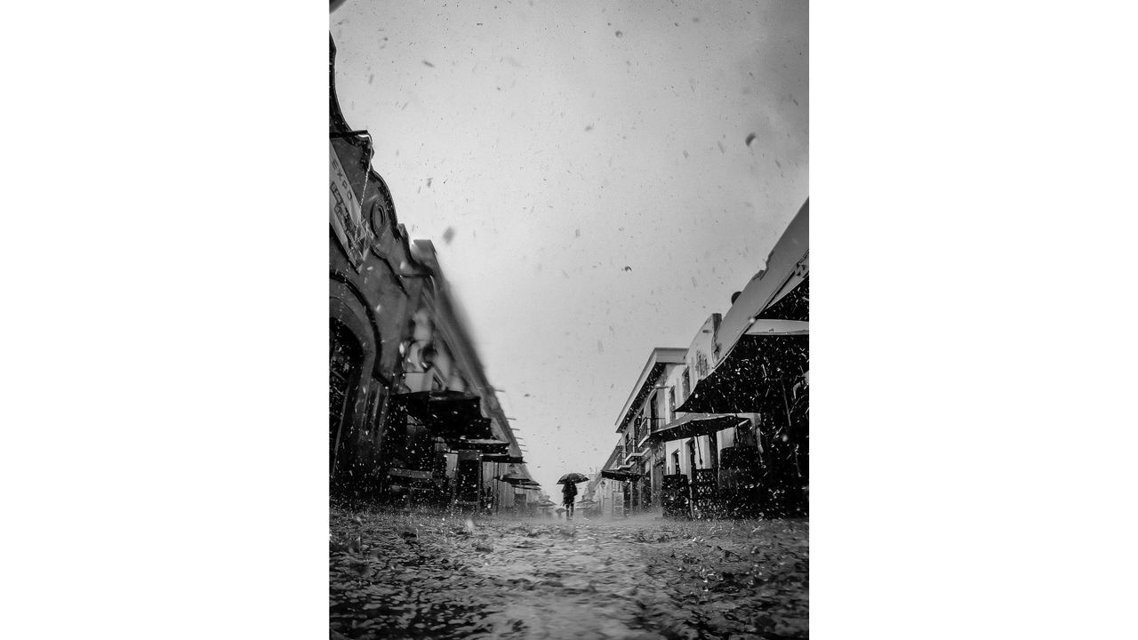 Black and white photo of a person with an umbrella walking down a rainy street between buildings with wet pavement and falling rain