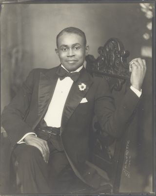 Black and white photo of Black man in bow tie and suit, smiling and seated in chair