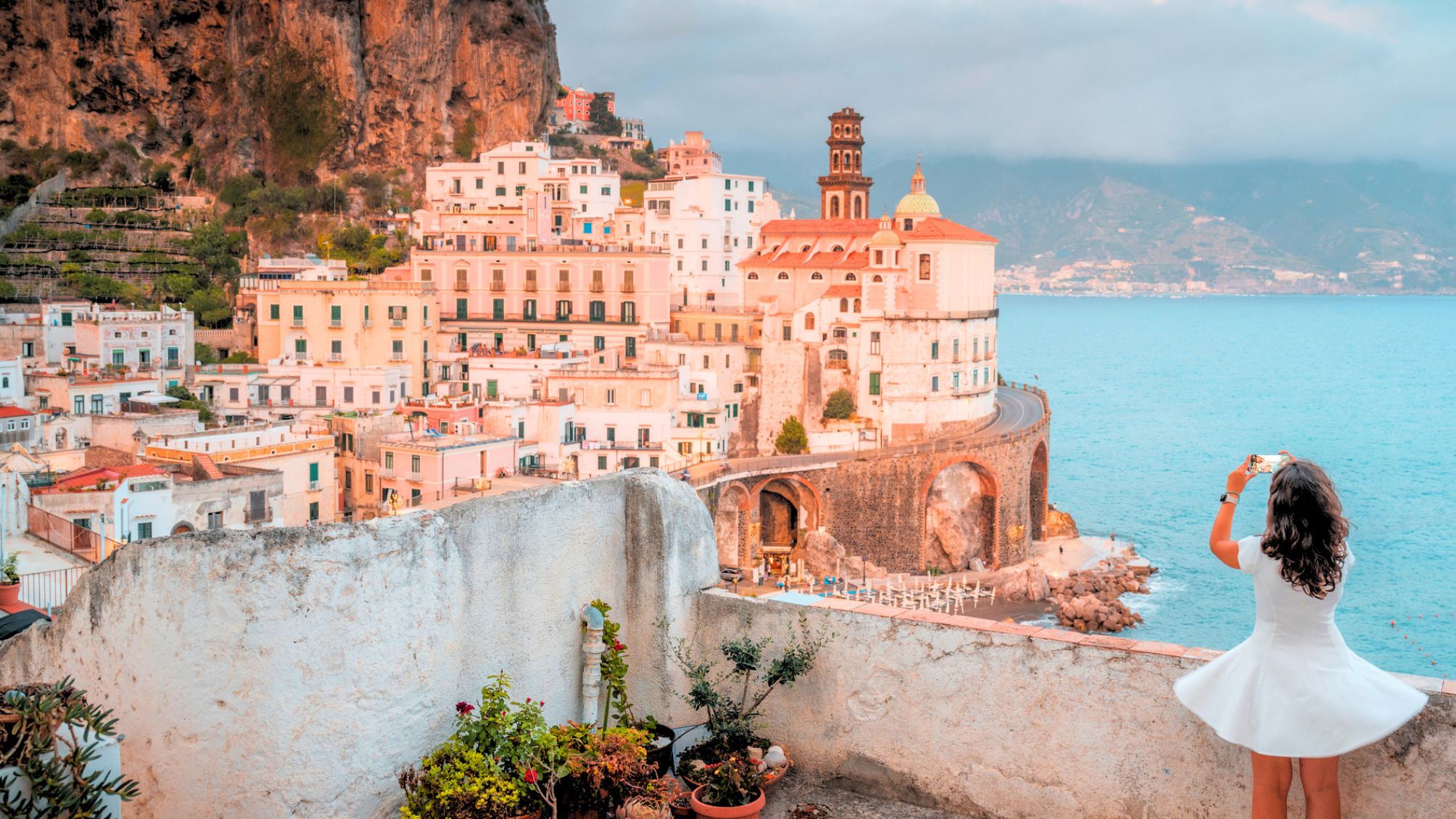 Woman in white dress photographing colorful coastal town on Amalfi coastline, Italy