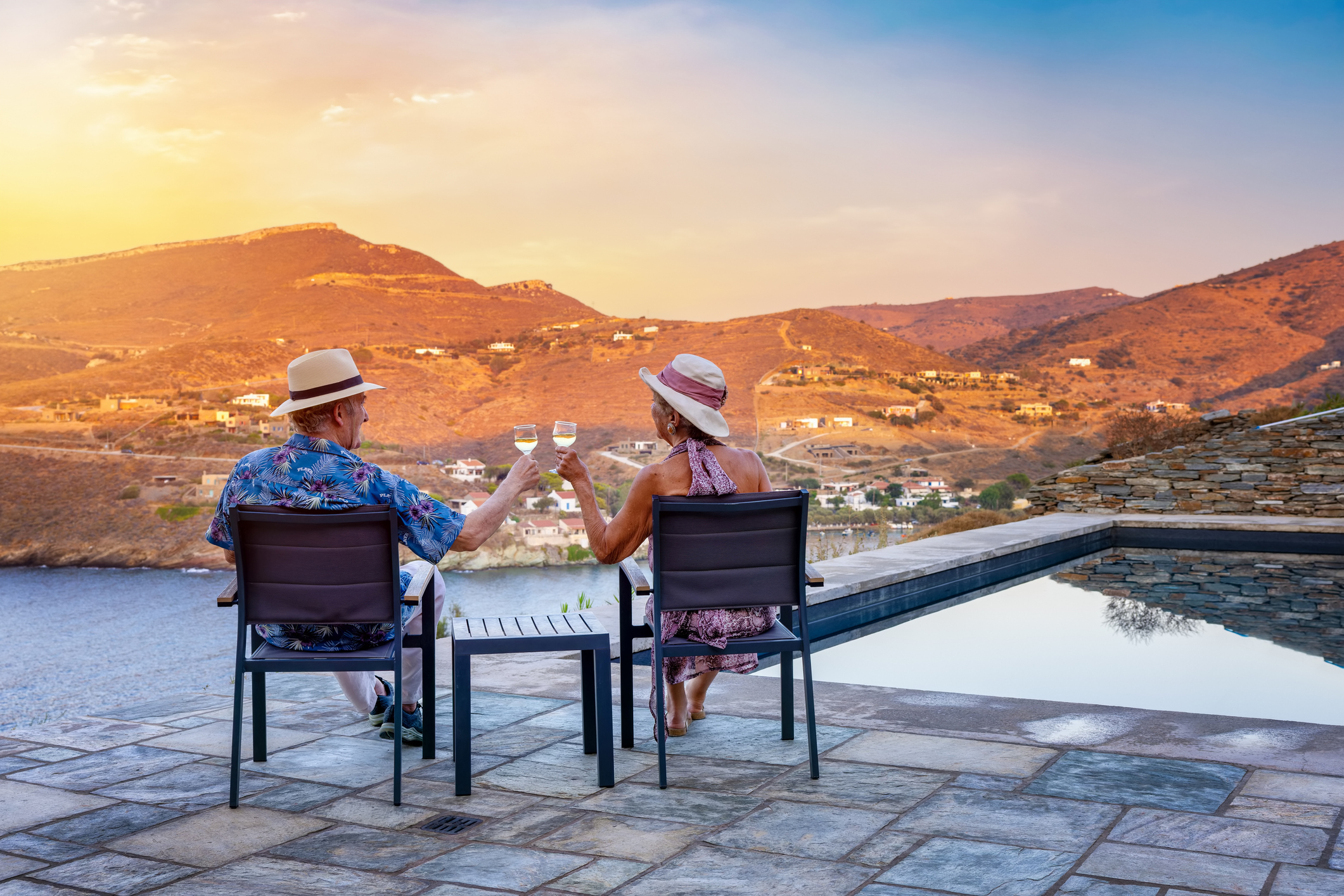 An older couple sits together by a swimming pool and enjoys the summer sunset in Greece