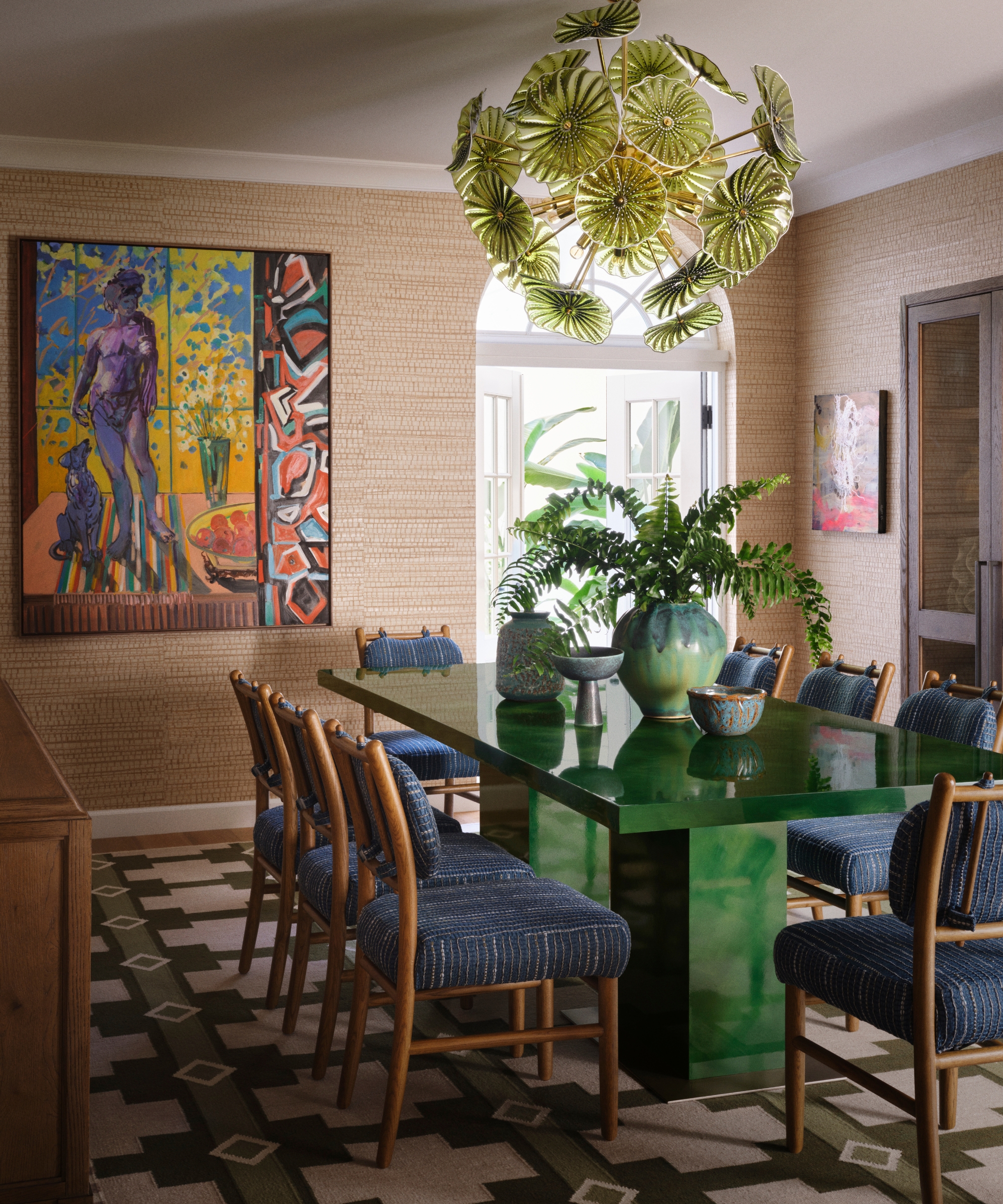 Dining room with glossy green table, blue upholstered chairs, leaf-form Murano chandelier, jute walls and contemporary art.