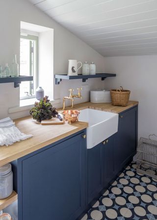 Small laundry room ideas under a sloped ceiling, with blue painted cabinetry and open shelving, wooden countertops and a blue and white tiled floor.
