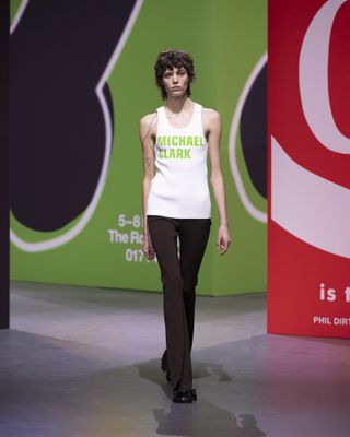 Woman on JW Anderson runway with coloured posters in background and tank top which reads 'Michael Clark'