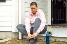 Woman tying up shoes on step outside a house, with a water bottle next to her