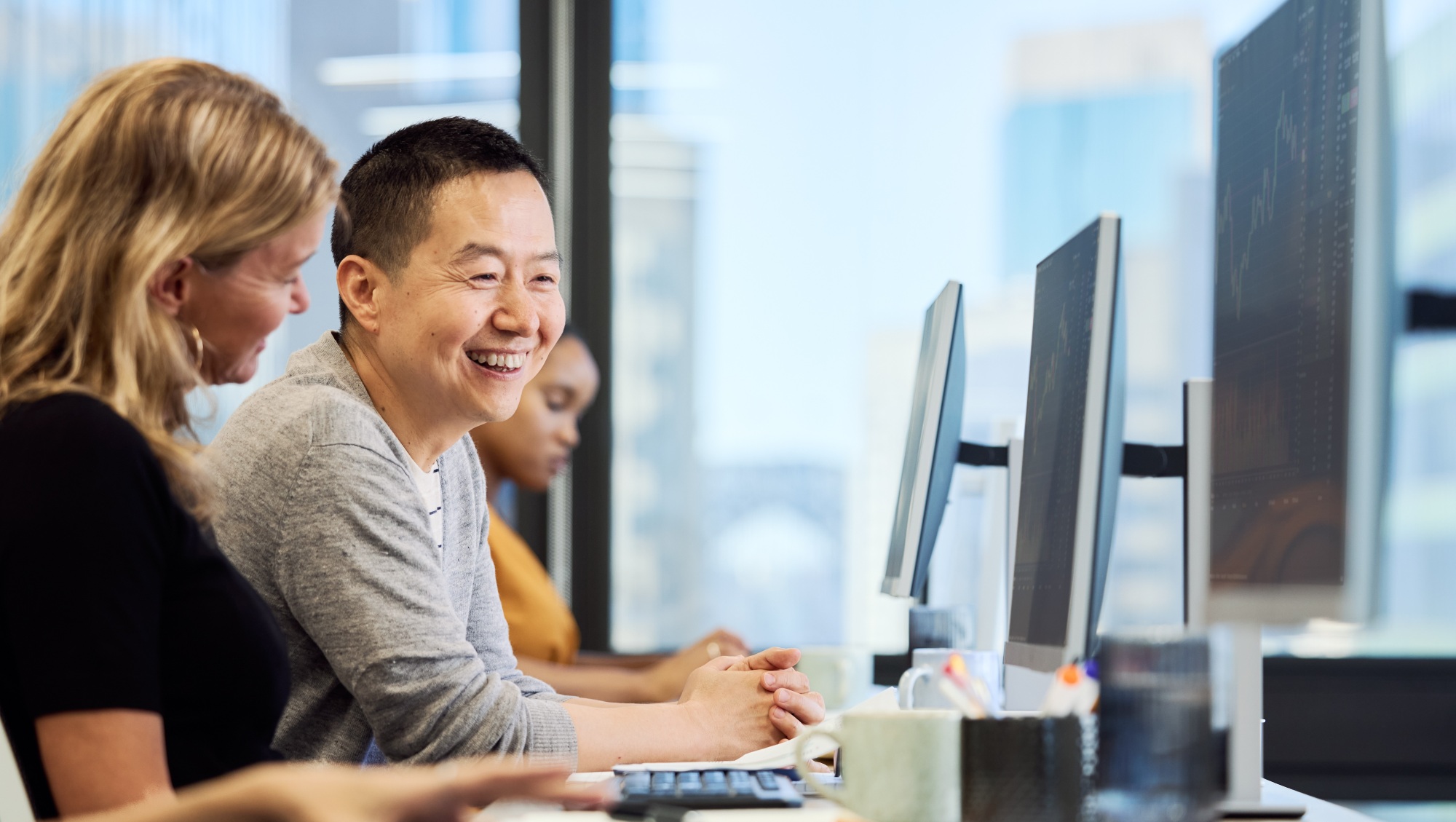 Happy male employee talking with colleague in office. Business people are sitting at desk. They are wearing smart casuals at work place.