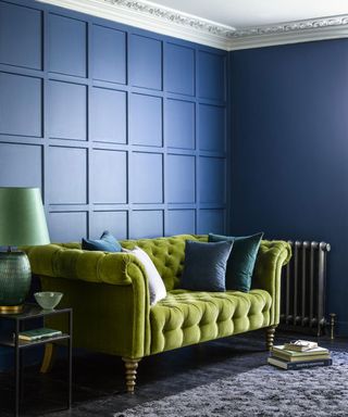 Blue painted living room with square paneling, green velvet two seater sofa, black floorboards and gray rug, black metallic side table with green table lamp