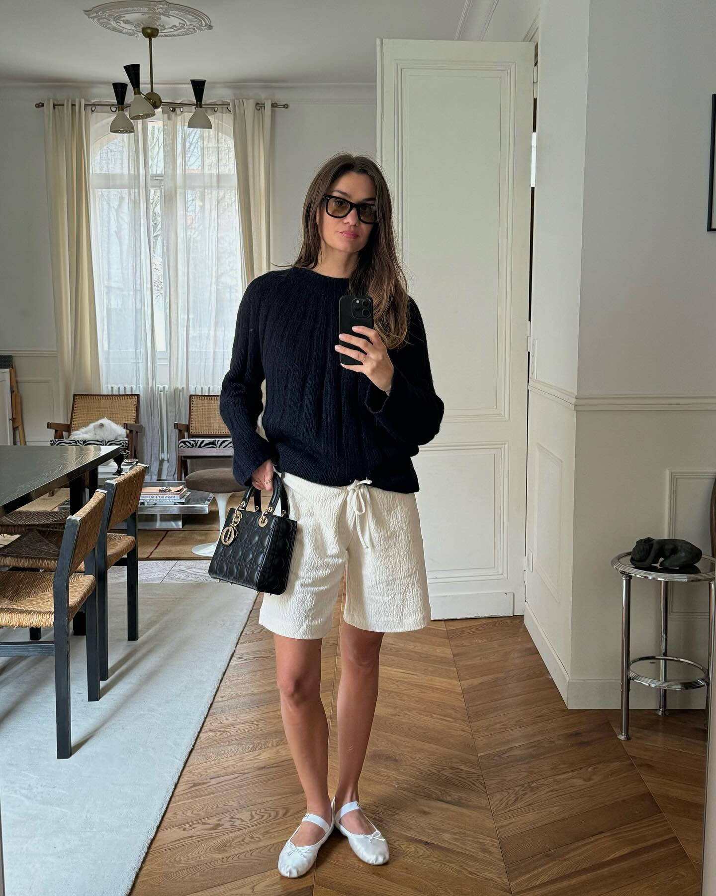 French fashion influencer Anne Laure Mais takes a mirror selfie wearing black sunglasses, a black sweater, mini Dior bag, textured drawstring white shorts, and white Miu Miu satin ballerina flats