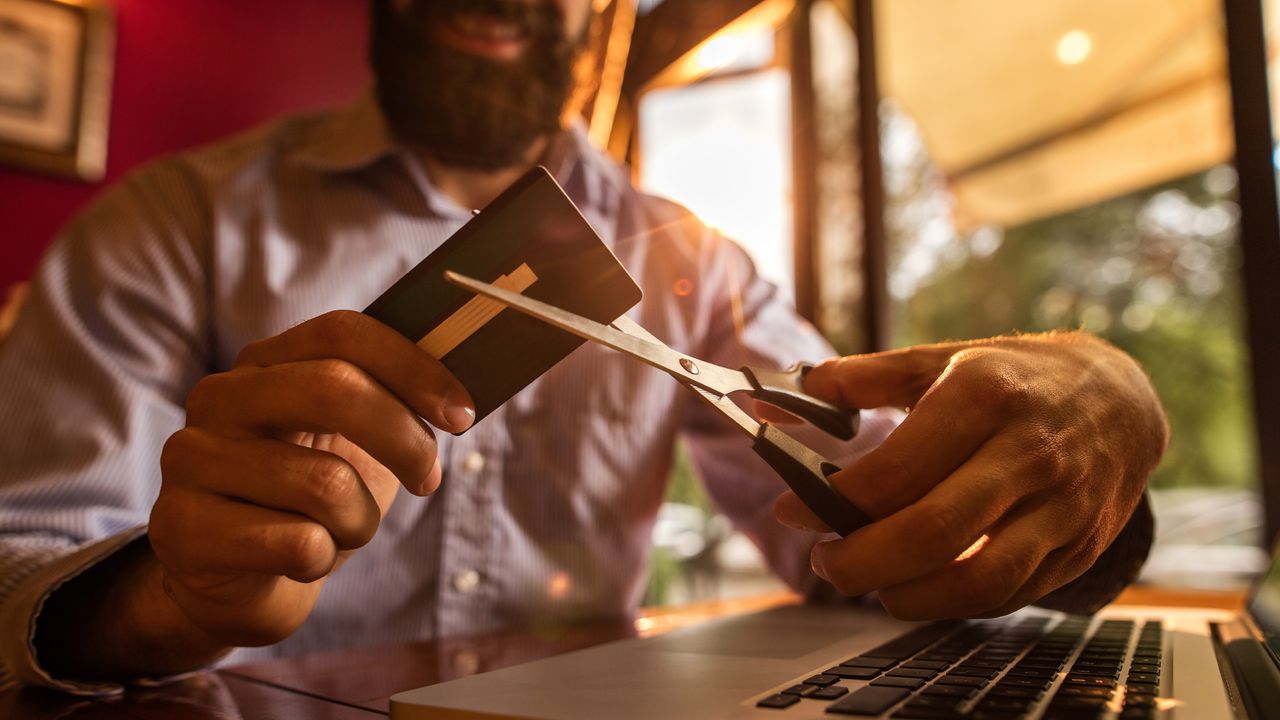 A smiling man cuts up a credit card while sitting at a desk in front of a laptop.