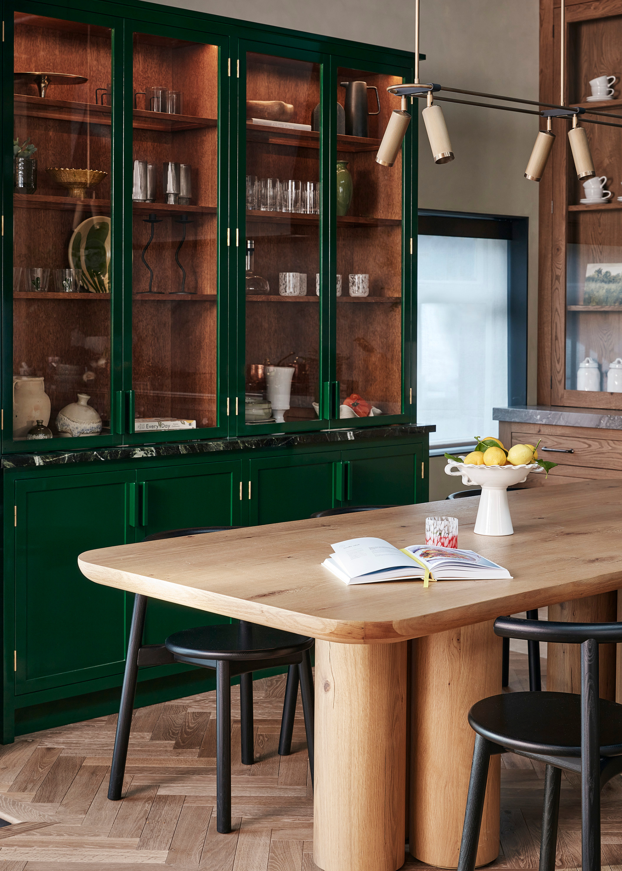 A modern kitchen with green gloss glass fronted kitchen cabinets filled with decorative glassware and tableware in front of a natural wood table with thick, circular legs with a fruit bowl and book open on top