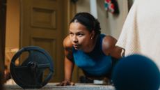 A woman performs push-ups next to a bed. She is on her toes and hands with her body held in a straight line. In front of her we see a barbell and small exercise ball.