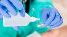 A person wearing turquoise scrubs and blue nitrile gloves pours clear water from a square plastic bottle onto a q-tip