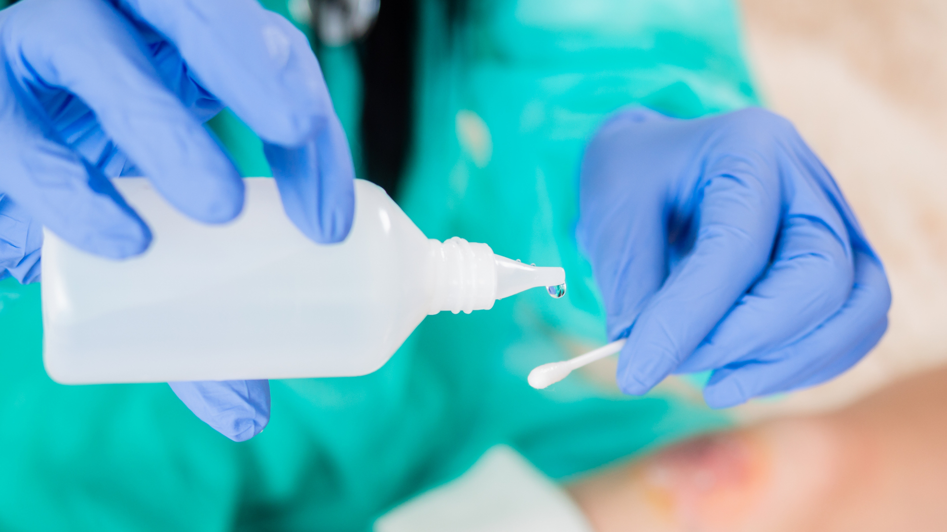 A person wearing turquoise scrubs and blue nitrile gloves pours clear water from a square plastic bottle onto a q-tip