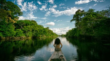 A girl with dark brown hair sits on the front of a wooden boat going through the Amazon rainforest in Peru