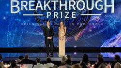 two people stand on stage in a glitzy ballroom awards show