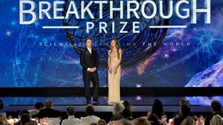 two people stand on stage in a glitzy ballroom awards show