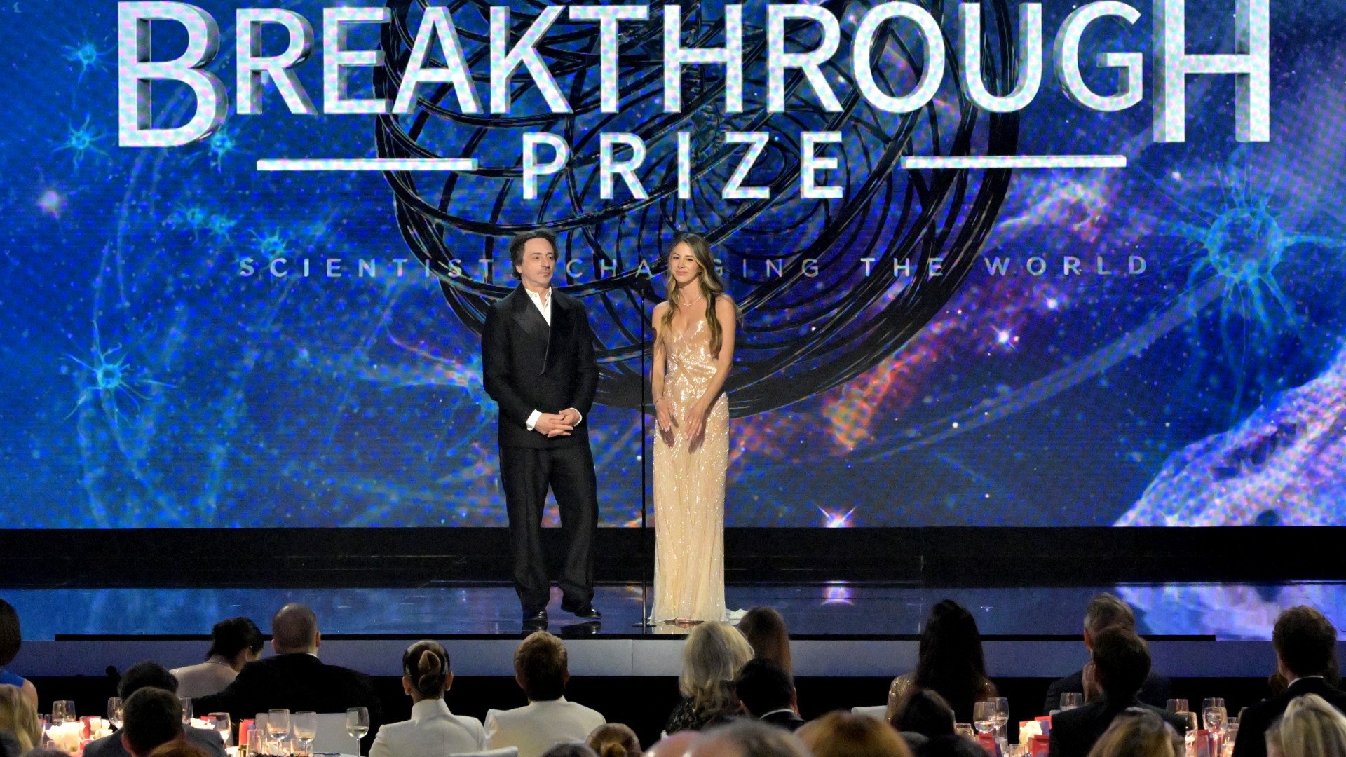 two people stand on stage in a glitzy ballroom awards show