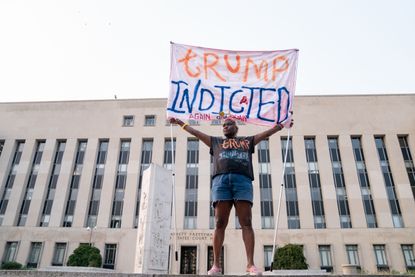 A person holds a sign reading "Trump Indicted" 