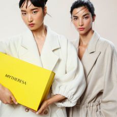 Two young woman in neutral wool coats with one holding yellow Mytheresa box