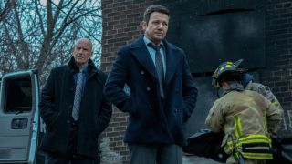 Hugh Dillon and Jeremy Rnner standing with their hands in their pockets in Season 4 of Mayor of Kingstown as firefighters walk behind them.