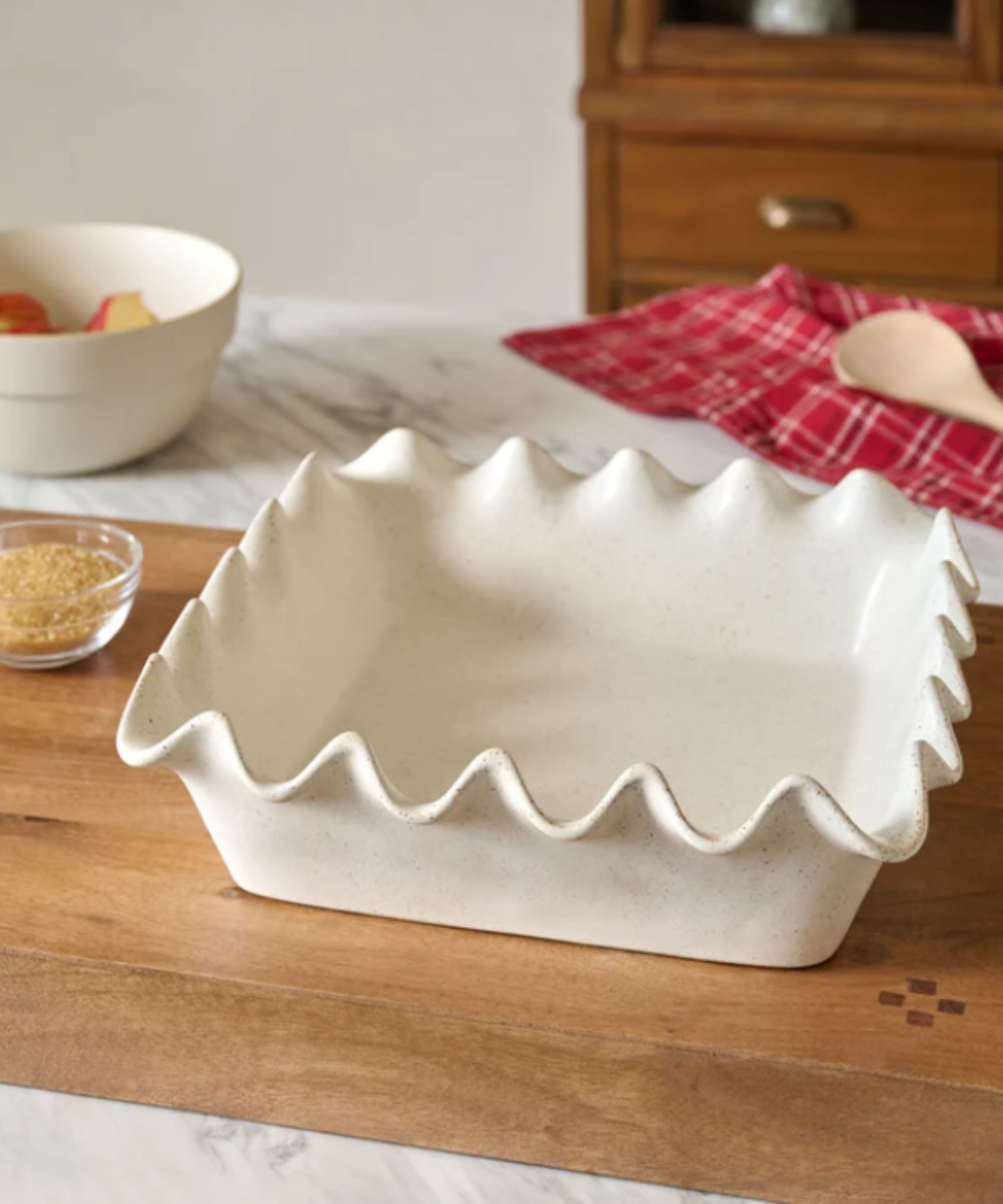 Square white scalloped pie dish on wooden board next to white ceramic bowl and red plaid dish towel