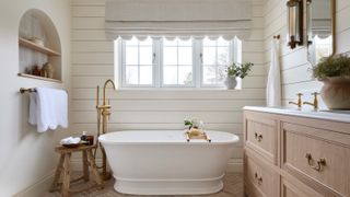 bathroom with white wall panelling, freestanding white bath, wooden vanity with gold taps, arched built in shelving unit and scalloped edged roman blind at window
