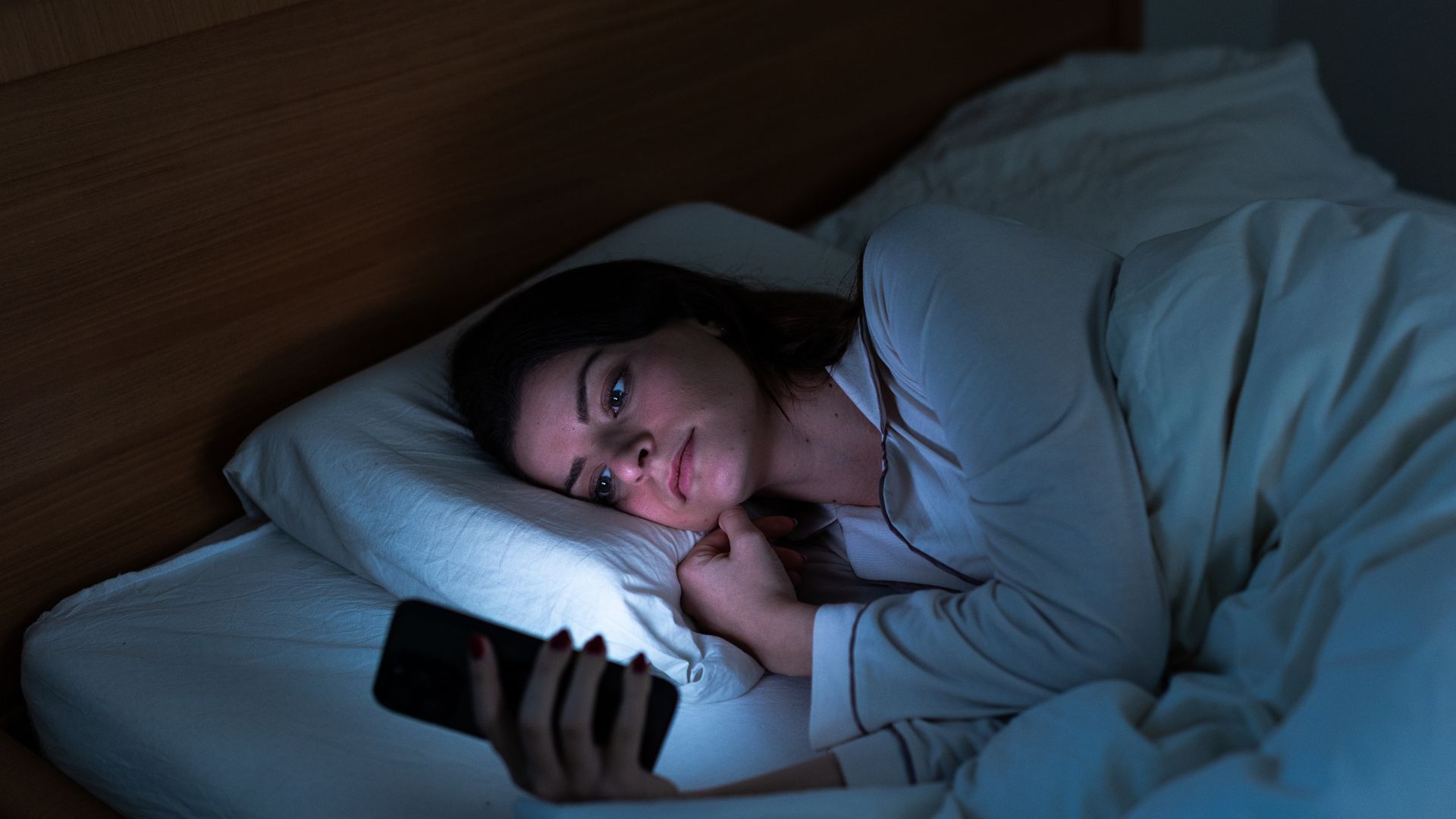 A woman lies in bed at night looking at her phone, she is illuminated by the light from the screen.