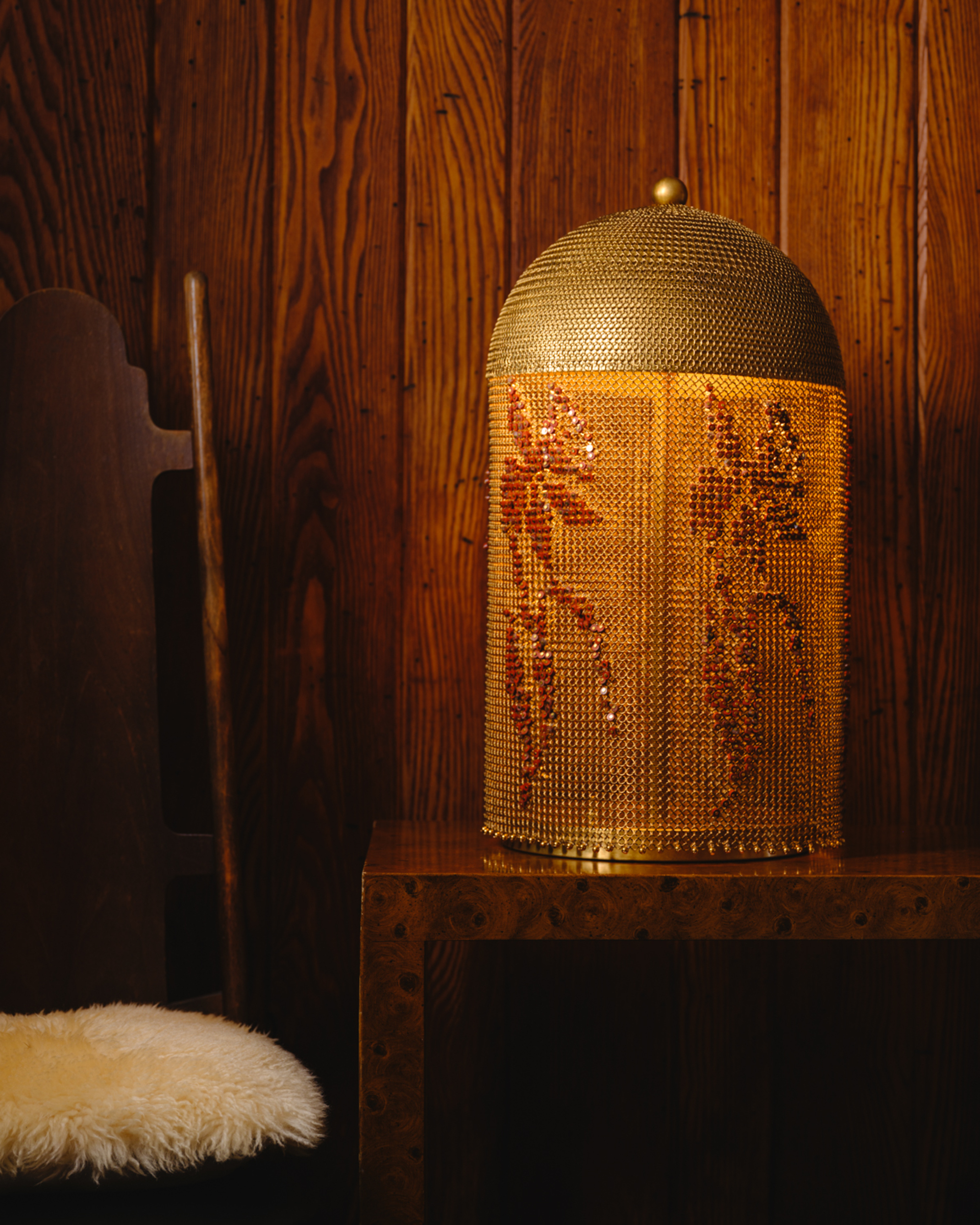 Close-up image of a lamp that is draped in chainmail and embroidered with beading. The lamp is on a wooden console table and the walls of the room are wooden. There is a wooden chair beside the table that has a white fur seat.