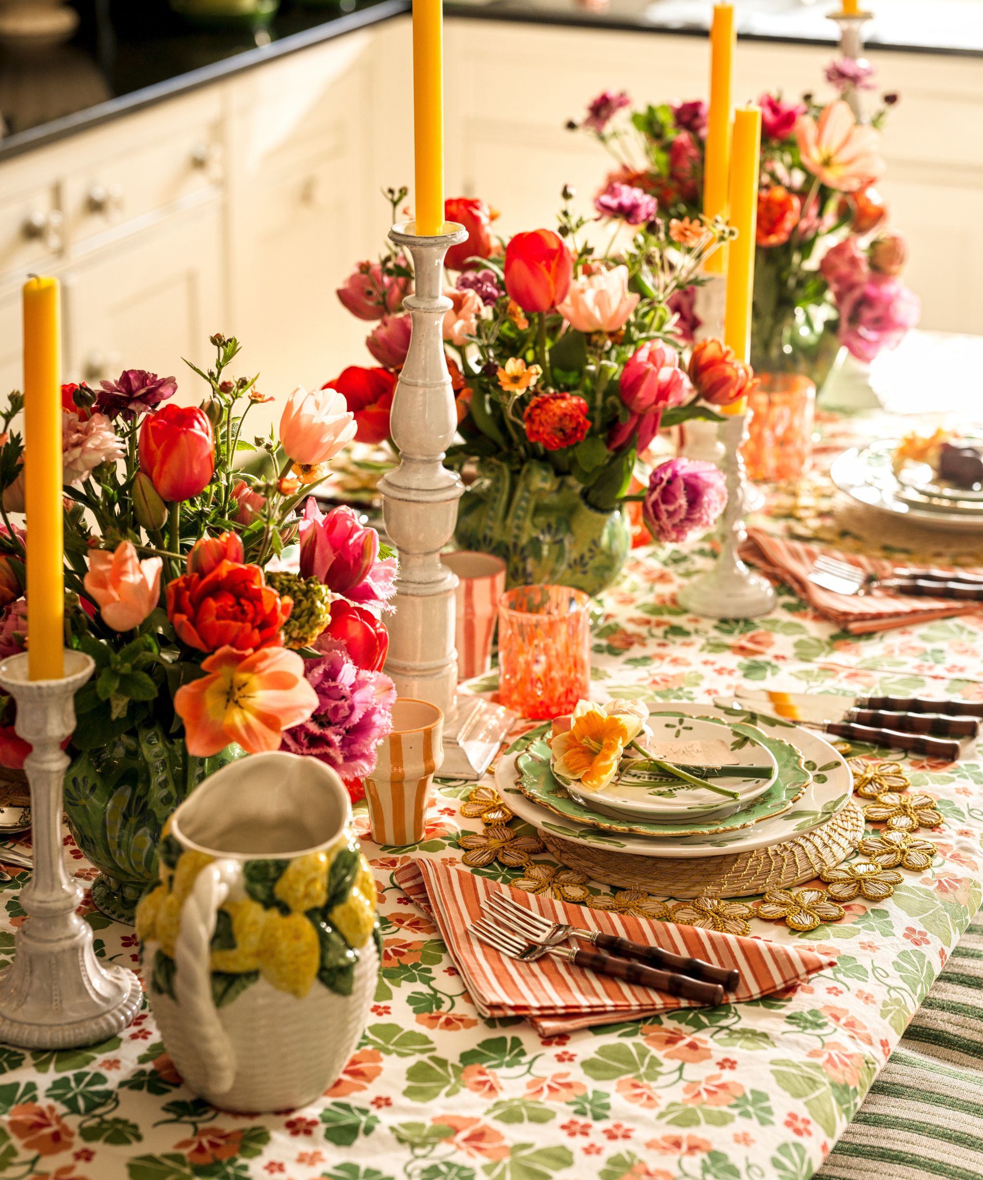 A high-angle view of a festive spring table set with vibrant tulip centerpieces, tall yellow taper candles in white holders, and green-rimmed plates topped with fresh daffodils
