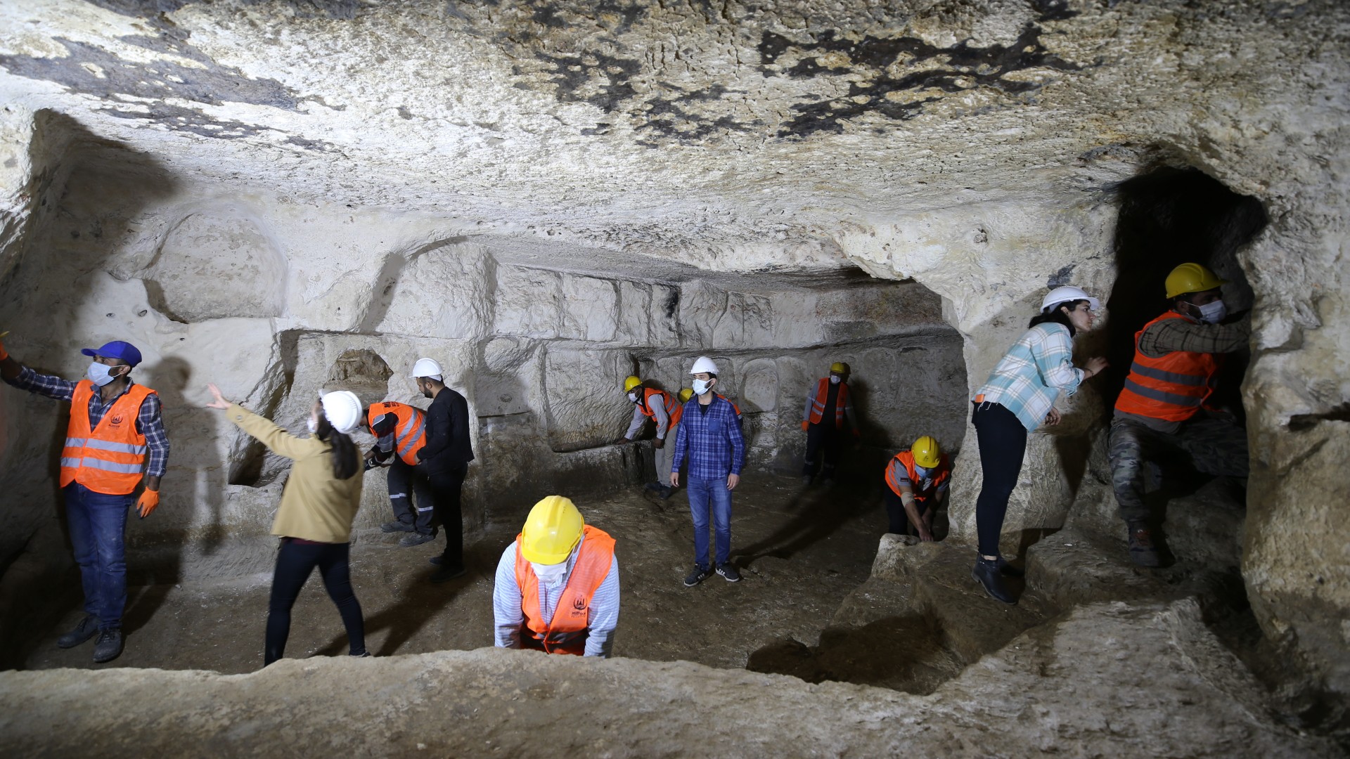 A team of 12 people who are wearing hard hats, masks and high visibility safety vests are exploring an underground cave thought to be a city.