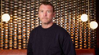 Sam Worthington in a black jumper standing in front of a gold background