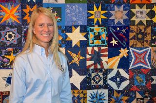 a smiling blond woman stands in front of a wall-mounted quilt