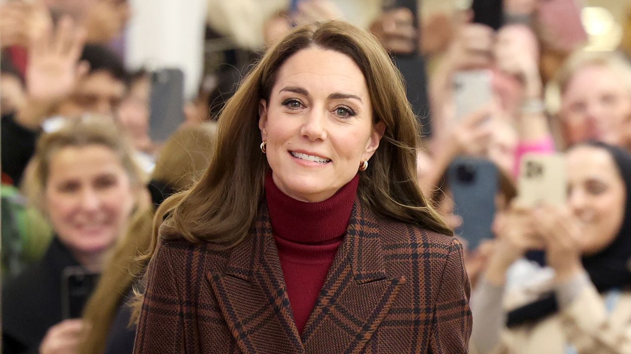 Kate Middleton has long brown hair and smiles while wearing a tartan coat and red sweater