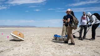 four people surround a small yellow-orange space capsule in a dry, flat desert lakebed.