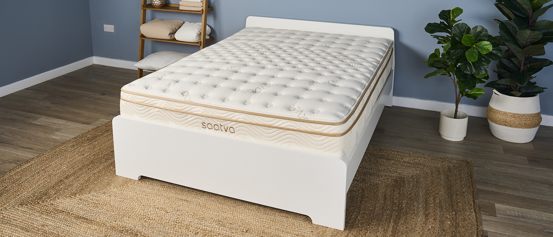 The Saatva Classic Luxury Innerspring Hybrid Mattress photographed on a white wooden bed frame in our blue colored Sleep Product Testing Lab. One one side of the mattress sit two tall green house plants, and on the other is a natural shelving unit containing pillows, weighted blankets, a sunrise alarm clock, and a variety of sleep tech.