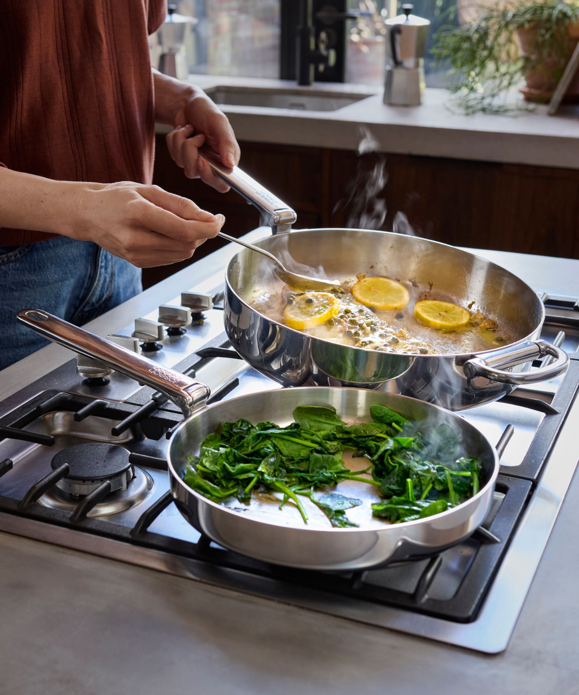 Two stainless steel frying pans being used to cook spinach and fry fish