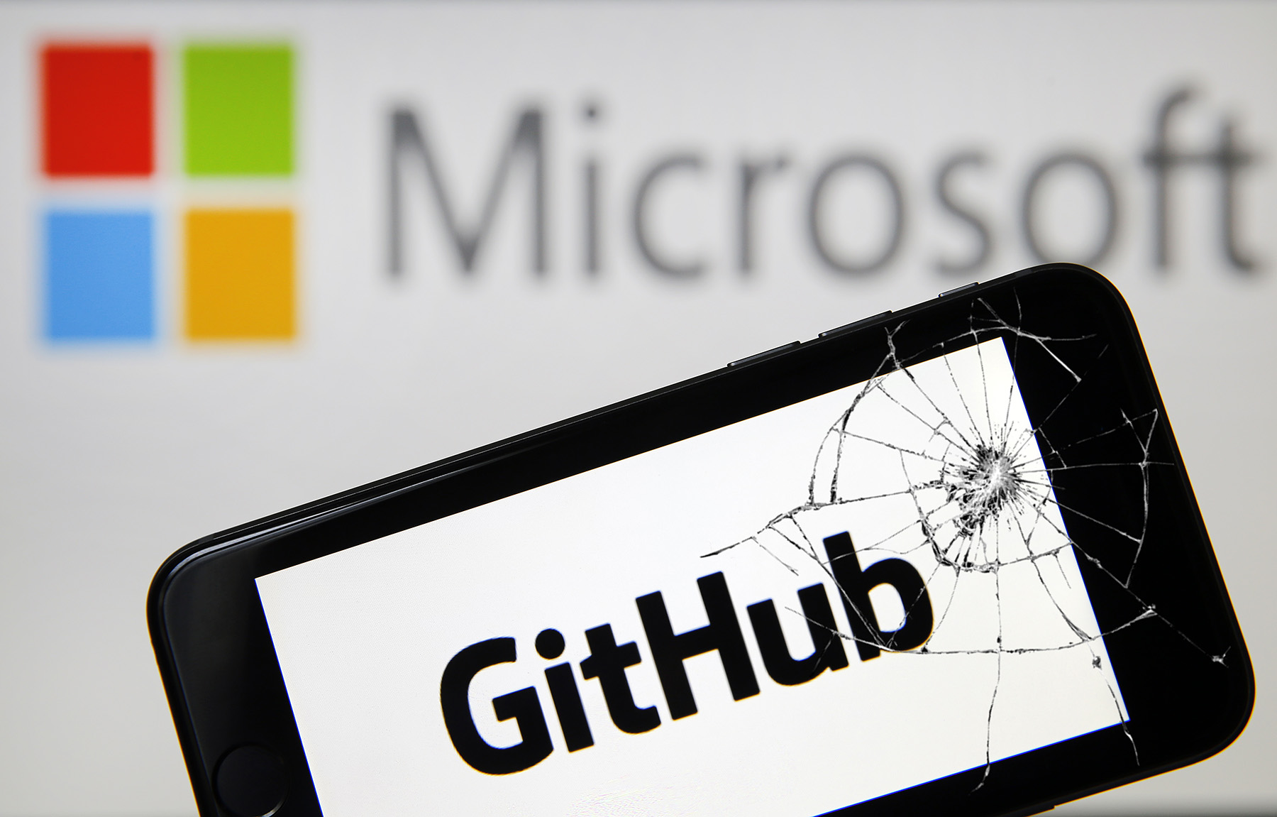 Github logo on a phone screen with a cracked display, with Microsoft's logo blurred in the background via depth of field. 