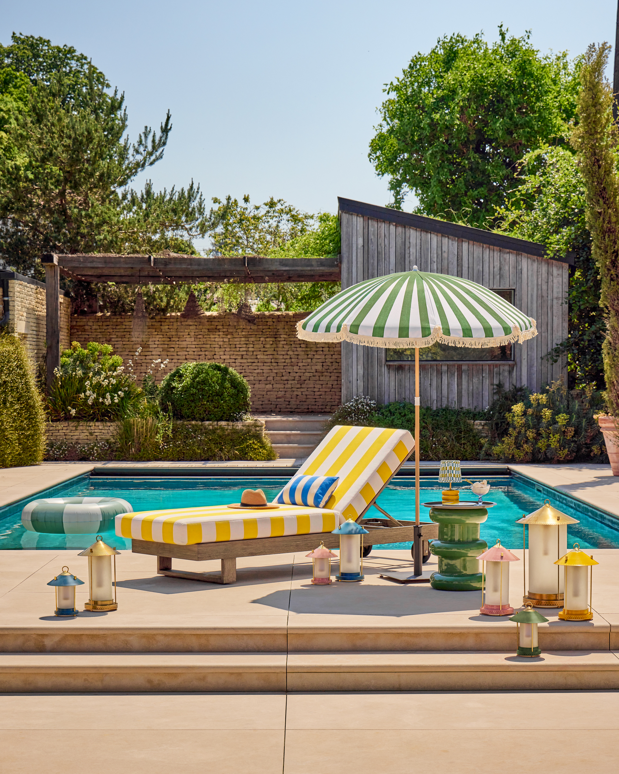 An outdoor pool and patio with a yellow striped sun lounger and a green striped parasol. There are lots of colorful outdoor lanterns around the seating area.
