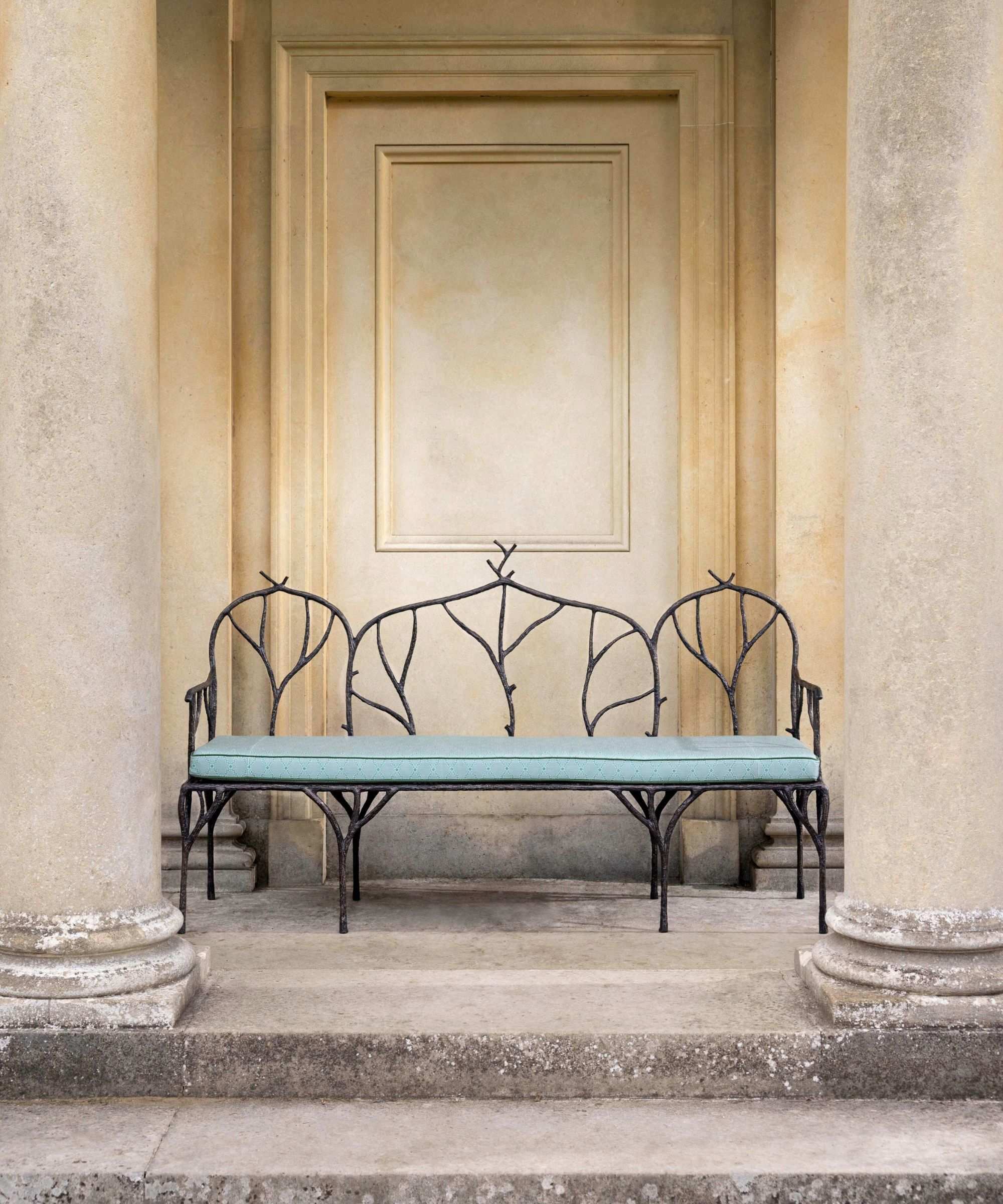 Stone pillars leading to an outdoor veranda with a three person bench with a back made from iron formed in the shapes of twigs and natural shapes