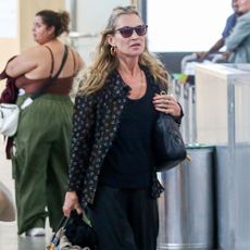 Kate Moss walks through the airport wearing black wide-leg jeans with a black vest top, a printed shirt and heeled boots.