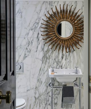 Bathroom with grey and white marbled floor and walls, white handbasin and toilet with starburst mirror on the wall.