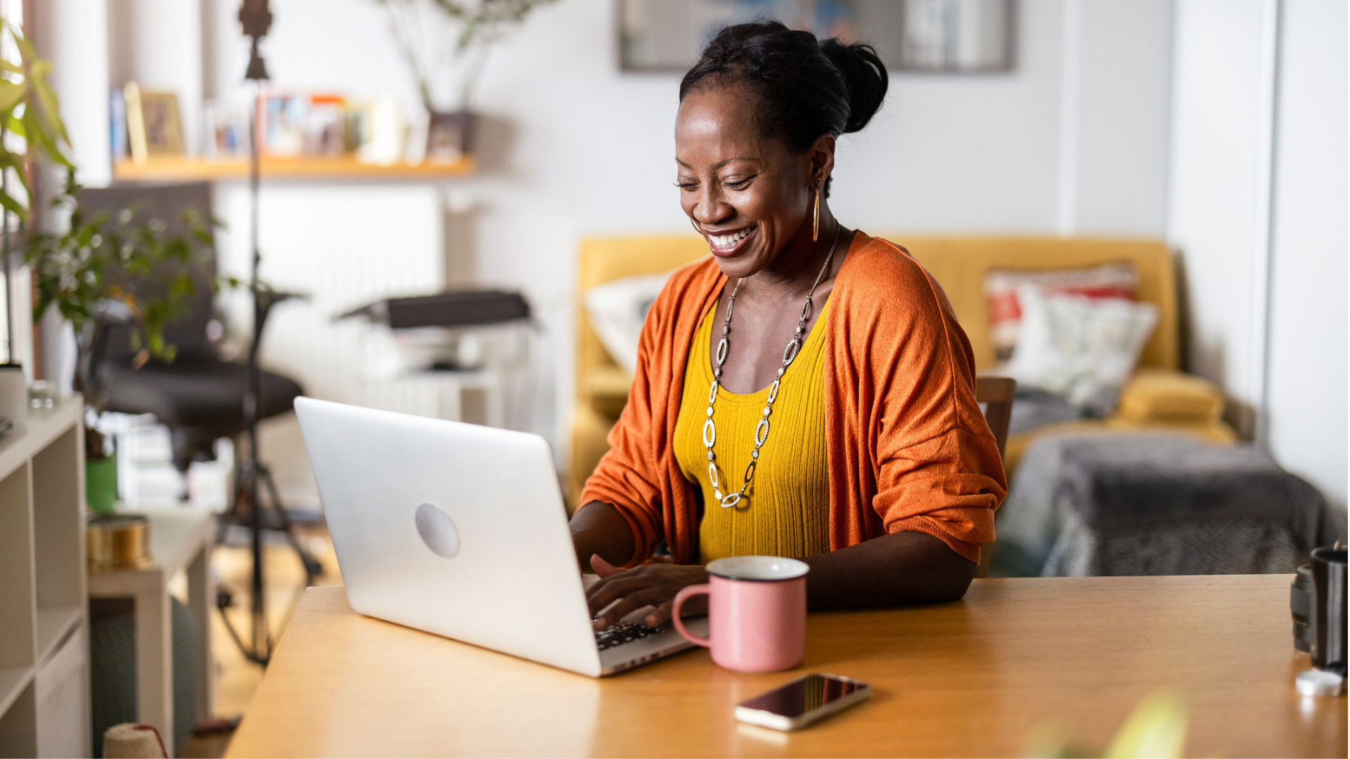 Smiling woman in a bright outfit browsing on a laptop in a stylishly-furnished room, with a mobile phone and a mug beside her.