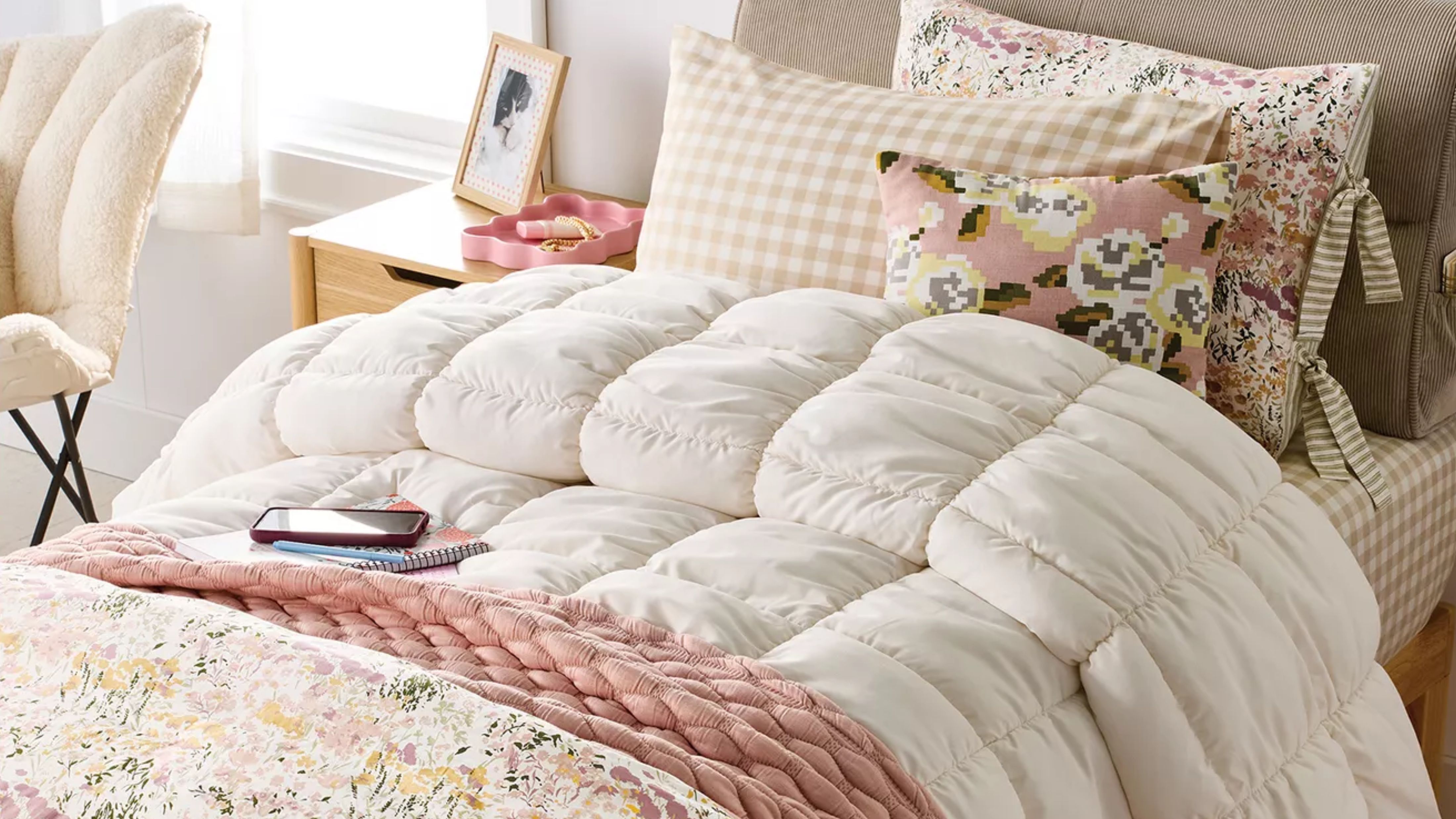 Target dorm essentials, including a cream tufted comforter, blush and floral throw blankets, and gingham sheets, all styled on a twin sized bed next to a window