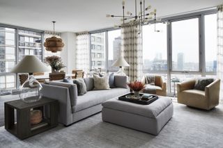 apartment living room with gray sectional and footstool, gray rugs, side tables, glass based lamps, focal point pendant lights, dining table in background