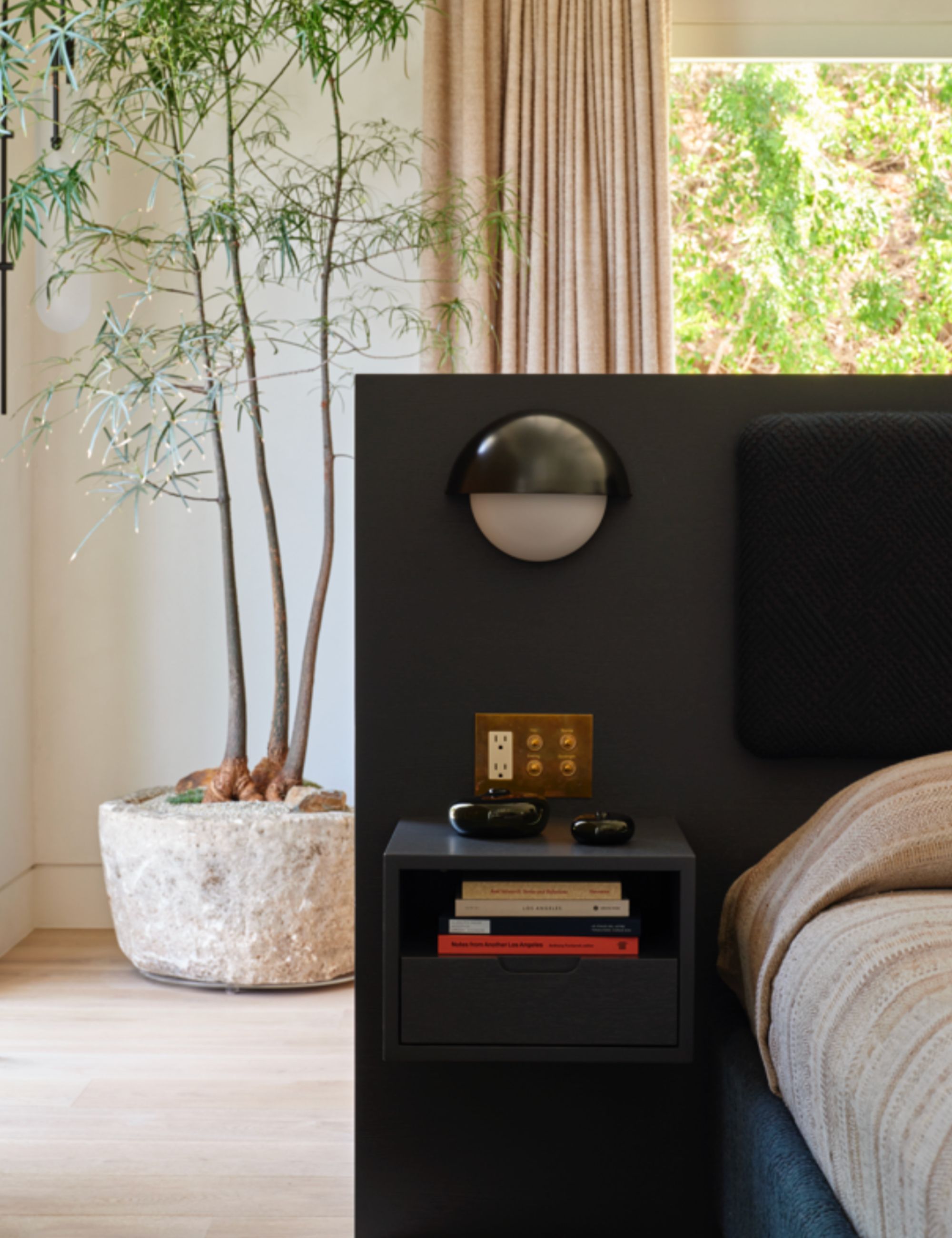 White bedroom with large tree in the corner, beige curtains, black headboard with round side light, floating side table with books, and a bed with beige linen and black velvet cushioning.