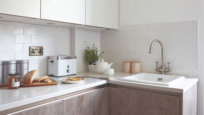 White kitchen with sink area and toaster, teapot, plant and sliced bread on chopping board on white worktop