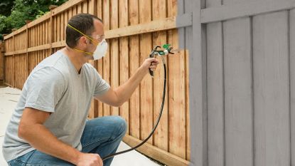 Man spraying fence with one of the best paint sprayer options and a grey paint, with a protective mask on